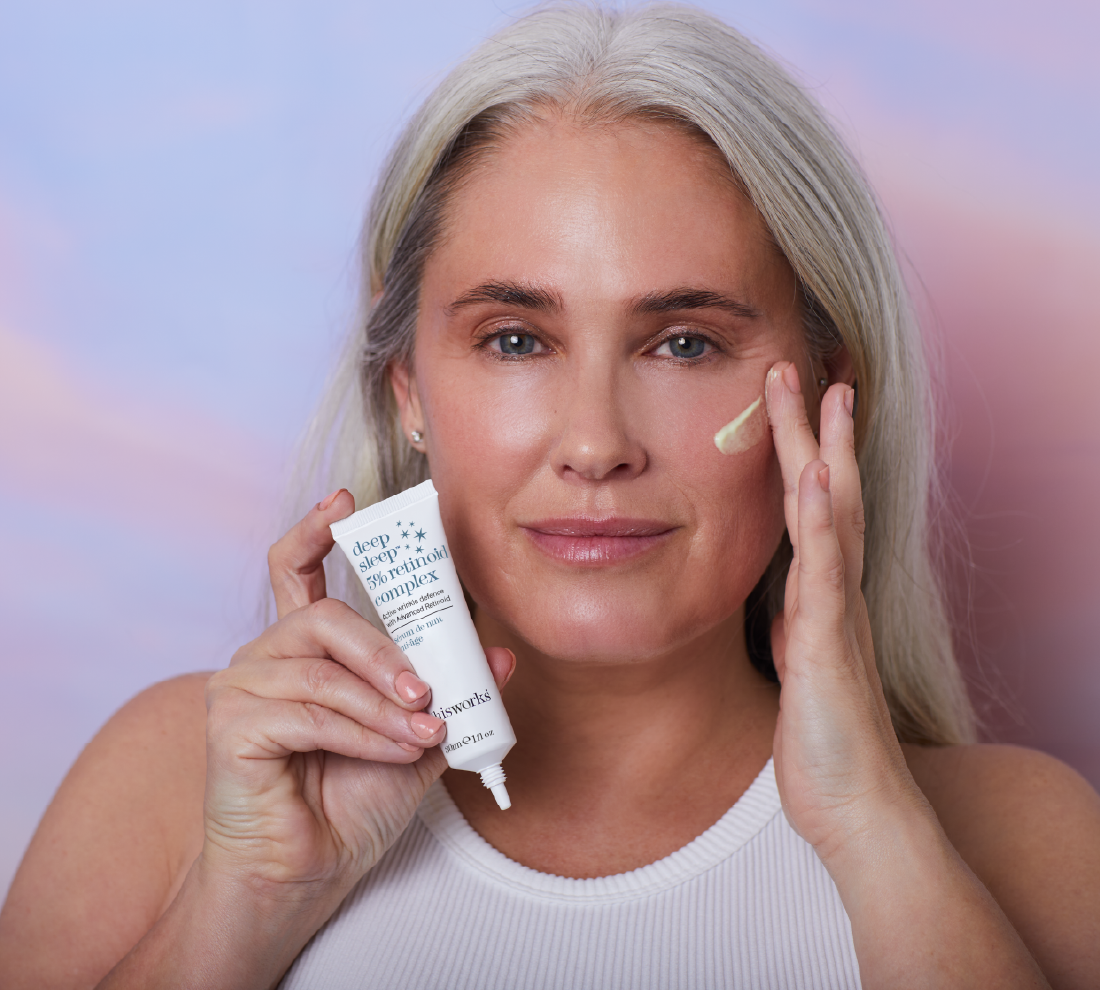 A woman applies the thisworks Deep Sleep 5% Retinoid Complex to her skin while holding the white tube of the product