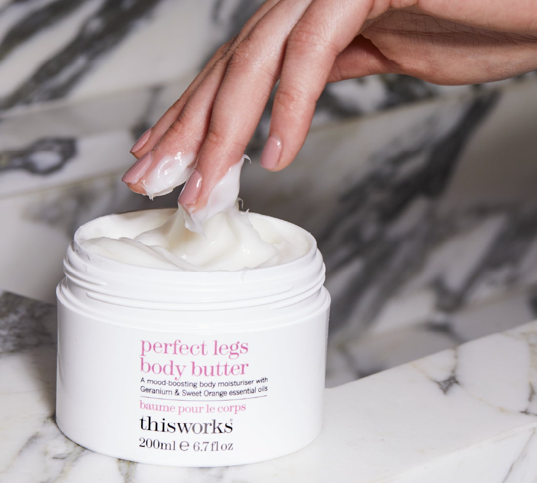 A close-up of a hand lifting a dollop of white body butter from a white jar labeled 'perfect legs body butter' by thisworks, set on a marble surface with a blurred marble background.
