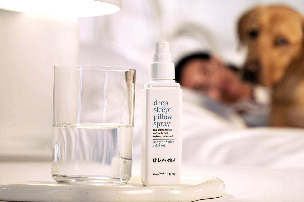 A bottle of thisworks Deep Sleep Pillow Spray stands next to a glass of water on a bedside table, with a blurred bedroom scene in the background showing a person sleeping in bed and a dog sitting nearby.