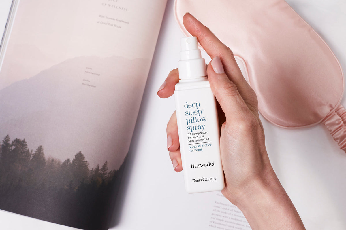 A hand holding a white bottle of thisworks Deep Sleep Pillow Spray next to a pink satin sleep mask and an open magazine.