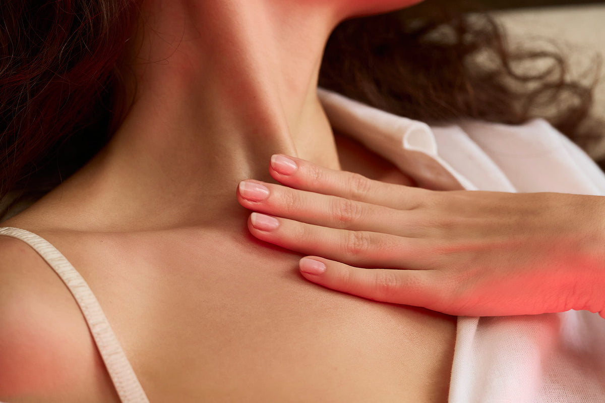 Close-up of a woman's neck and upper chest with a hand resting on her collarbone, under warm red lighting.