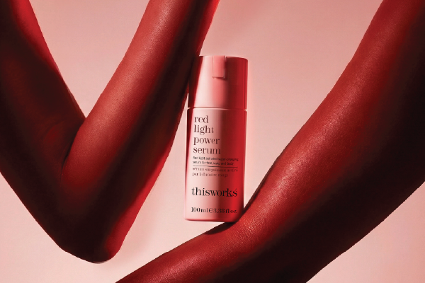 A bottle of thisworks red light power serum centered against a pink background, framed by two  arms extending from opposite sides.