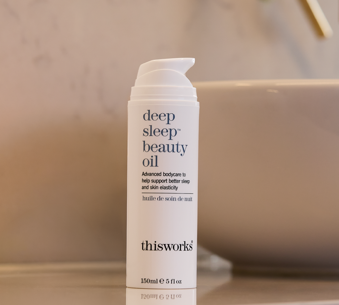 A 150ml white pump bottle of thisworks Deep Sleep Beauty Oil standing on a countertop with a softly blurred bathroom sink and wall in the background.