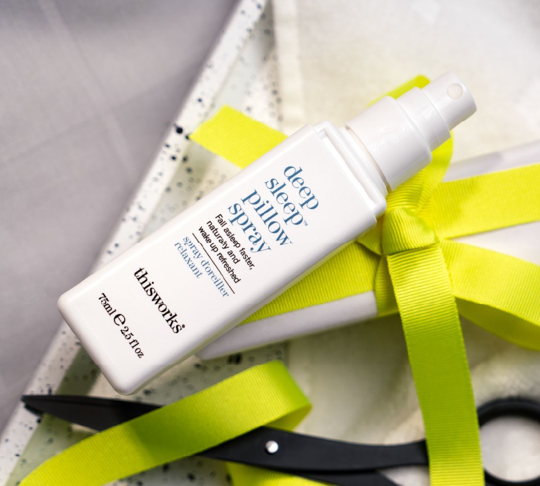 A white spray bottle of thisworks Deep Sleep Pillow Spray, styled diagonally with a bright yellow ribbon on a textured white and gray background.