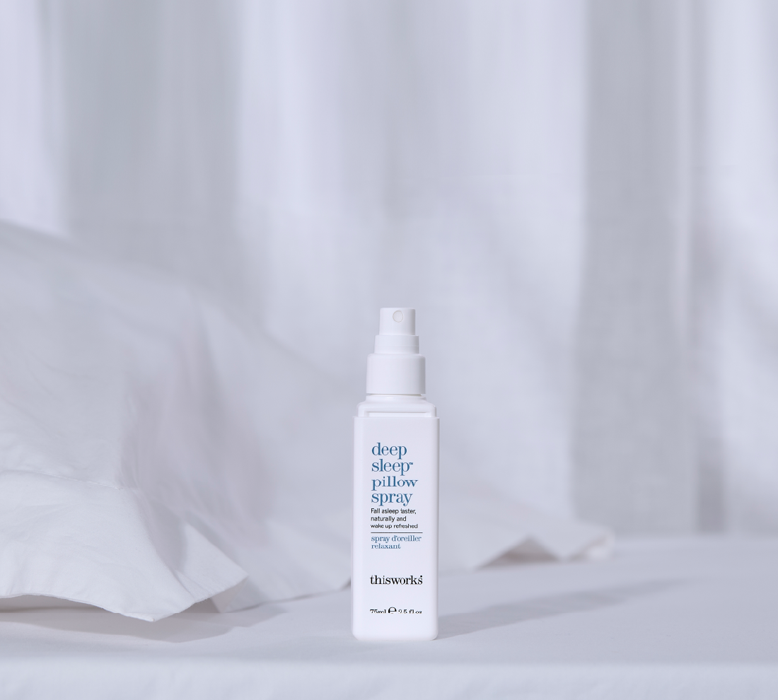 A white spray bottle of This Works Deep Sleep Pillow Spray standing on a white surface against a soft white draped fabric background.