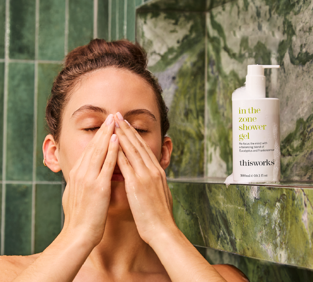 A woman standing beside a thisworks in the zone shower gel, inhaling the scent while in the shower