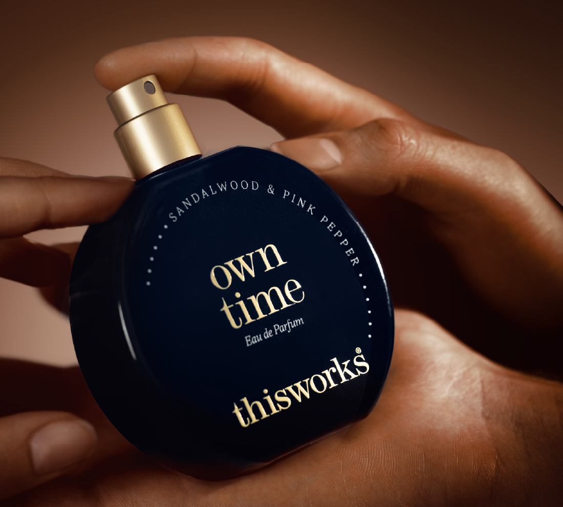A hand holds a dark navy perfume bottle with a gold spray top. The bottle is labeled 'own time Eau de Parfum' and 'thisworks' with 'SANDALWOOD & PINK PEPPER' written above.