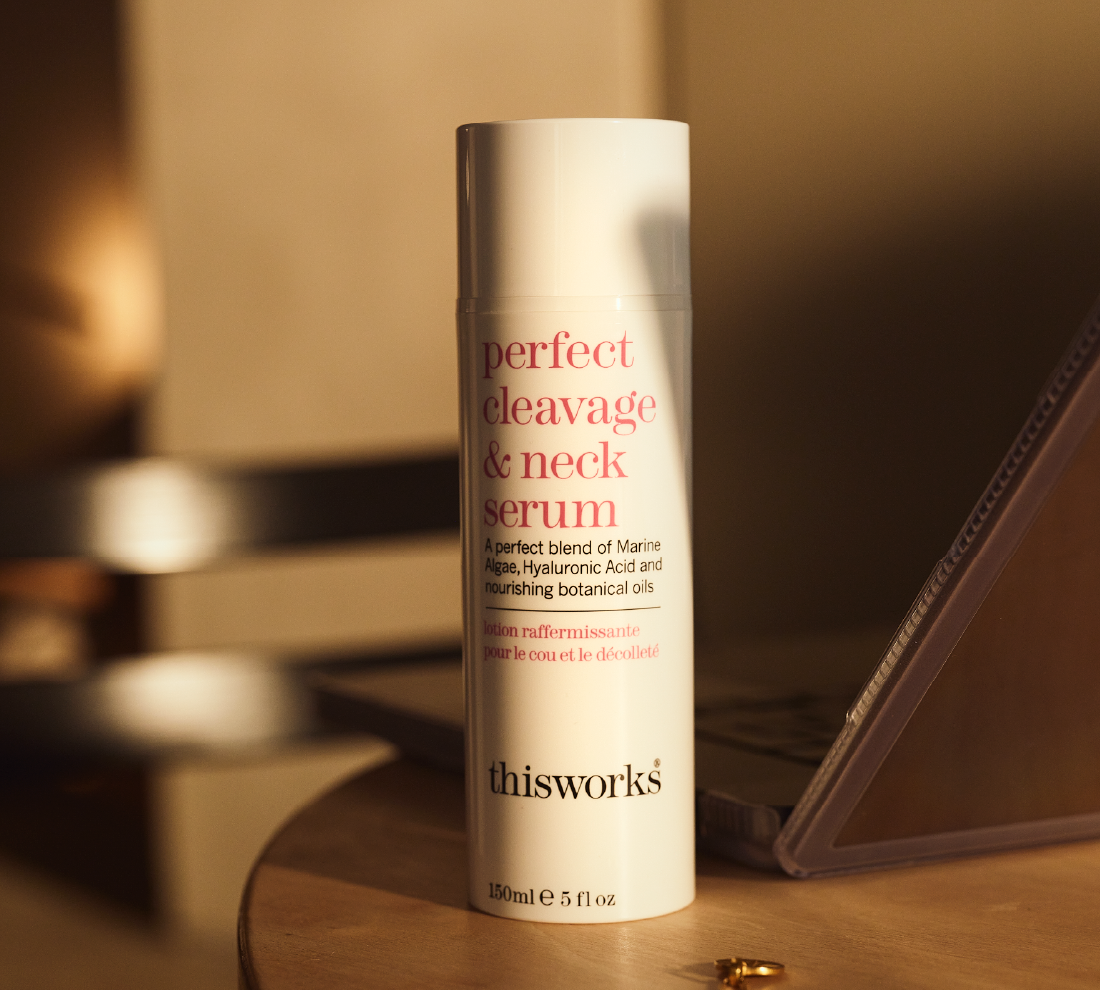 A white bottle labeled 'perfect cleavage & neck serum' by thisworks, standing on a wooden surface with a softly blurred warm-toned interior background.