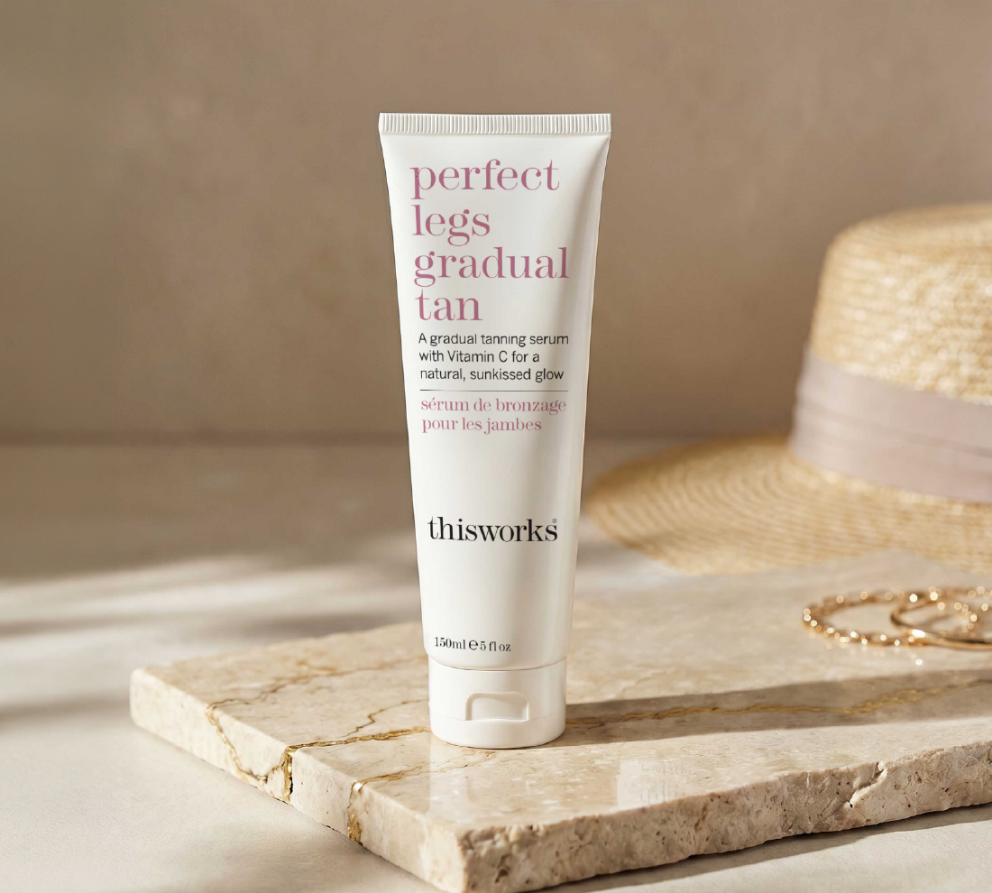A white tube of thisworks Perfect Legs Gradual Tan serum stands on a light stone tray, with a woven straw hat and gold jewelry softly blurred in the background against a warm beige wall.