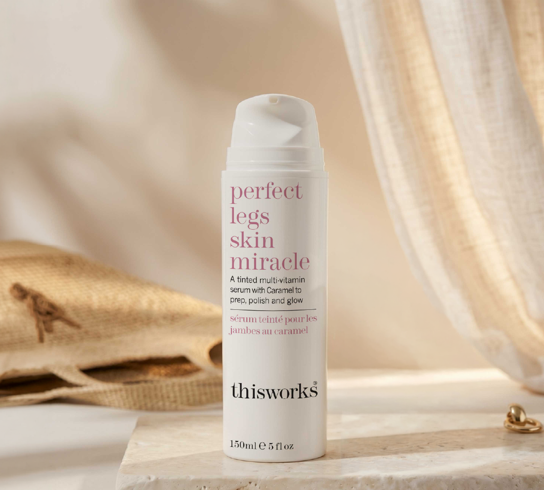 A white pump bottle of thisworks 'perfect legs skin miracle' serum, styled on a marble surface with a linen bag and draped fabric in the background.