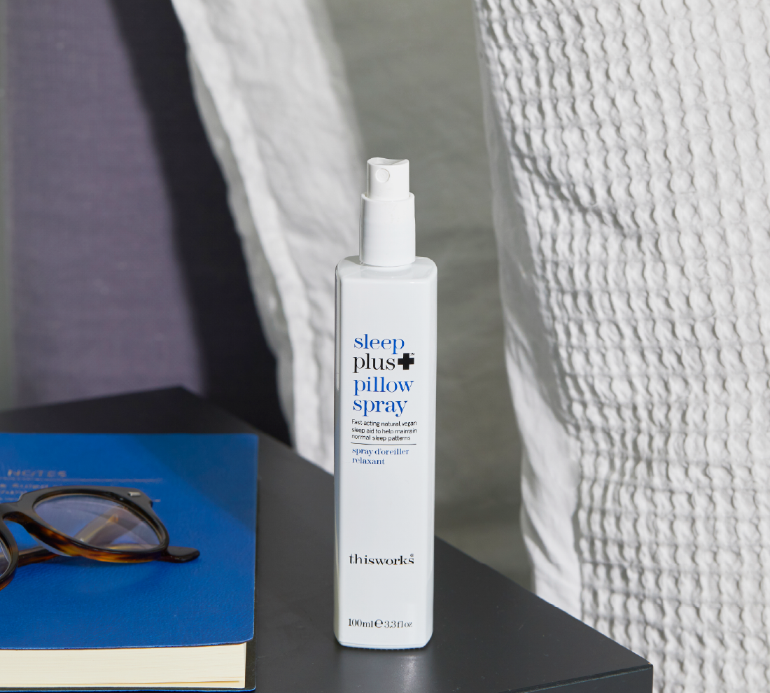 A bottle of Thisworks Sleep Plus Pillow Spray stands on a dark tabletop next to a blue book with glasses resting on it, with a white textured pillow or bedding in the background.