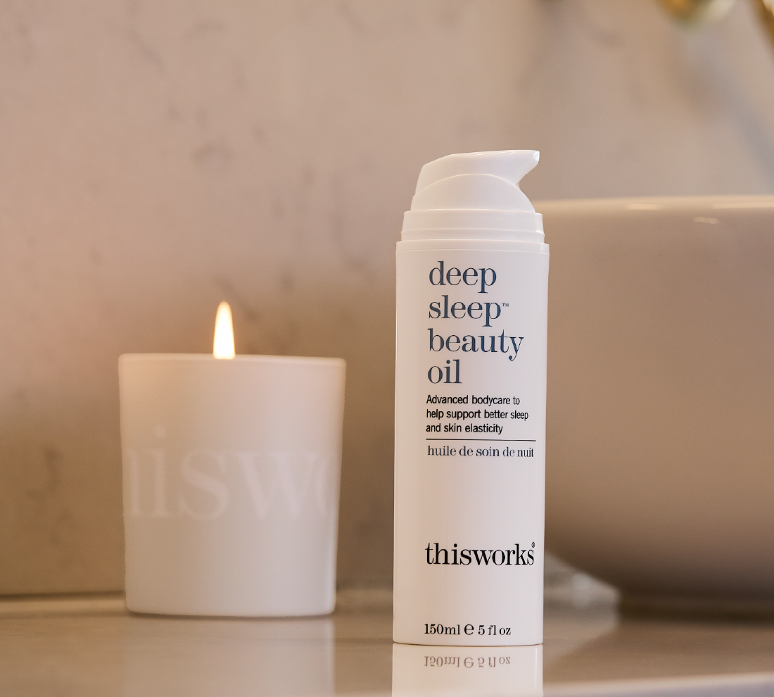A white pump bottle of thisworks deep sleep beauty oil stands on a bathroom counter, with a lit candle and a soft-focus sink in the background.