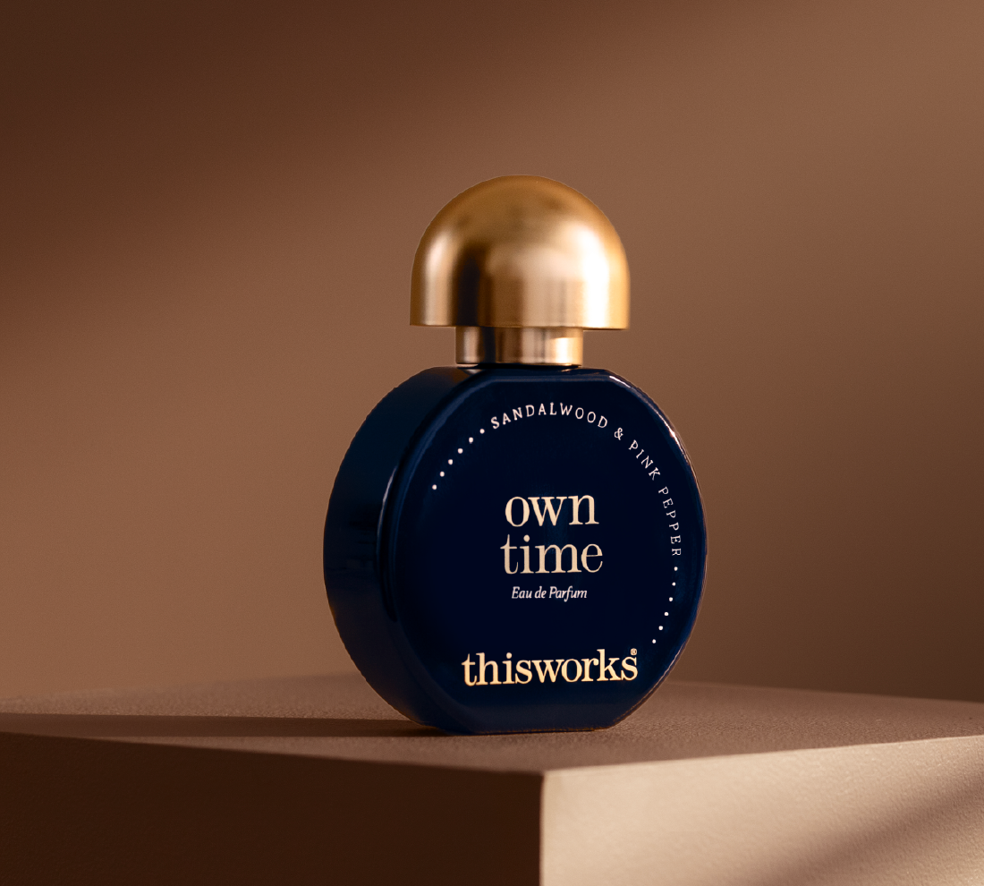 A bottle of thisworks Own Time Eau de Parfum with a dark blue body and a metallic gold cap, displayed on a matching gold pedestal against a warm brown background.
