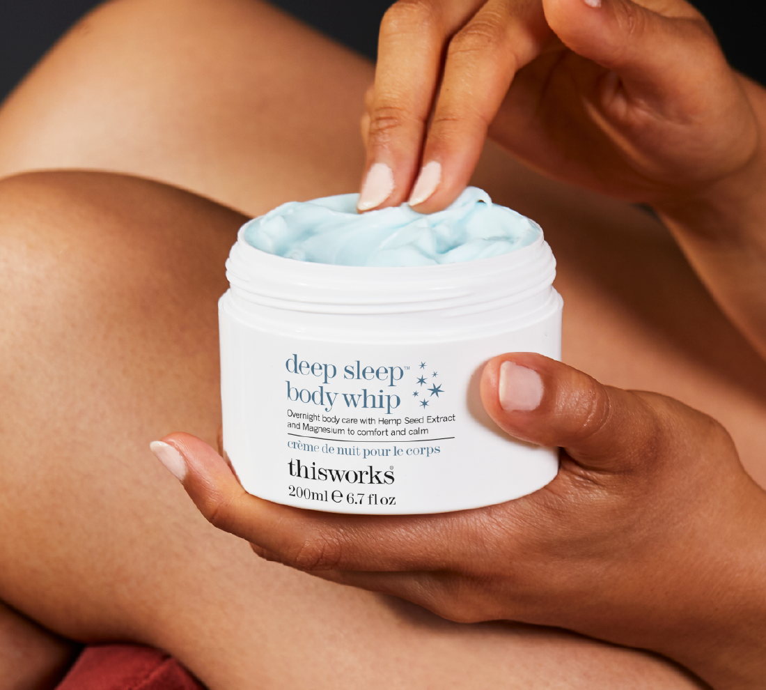 A person holds an open white jar of This Works Deep Sleep Body Whip, showing the light blue cream inside. The person's hands and legs are visible in a warm, close-up shot.