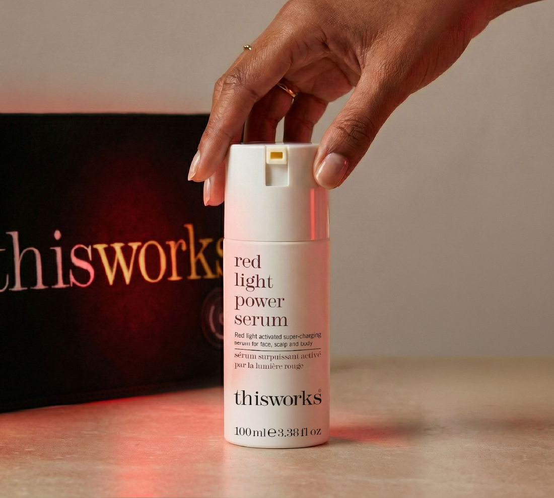 A hand holds a bottle of thisworks Red Light Power Serum against a warm beige background with the red light body wrap in the background.